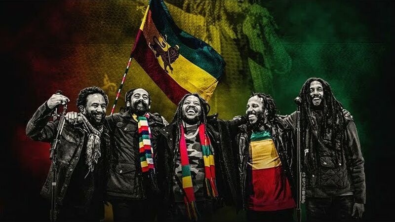 The Marley Brothers stand arm in arm in front of a background featuring the Jamaican flag. They wear the green, yellow and orange colors of the flag.