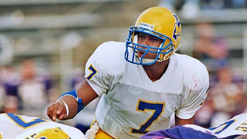 In this image, Kerry Joseph, a quarterback for McNeese State University from 1992-1995 and member of the Louisiana Sports Hall of Fame, is seen in action on the football field. Wearing a blue and yellow helmet with a face mask, a white jersey with the number 7, and a blue armband, he appears focused and ready to lead the play. His stance and gaze suggest he is observing the field, possibly preparing to make a pass or direct his team. The blurred background hints at a stadium setting with spectators.