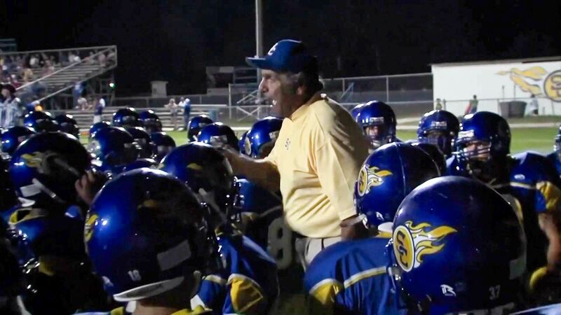 Frank Monica is seen passionately coaching a football team during a nighttime game. He is wearing a light yellow shirt and a blue cap, addressing a huddle of players in blue and yellow helmets featuring a flame logo. The players listen intently as he gestures, providing guidance and motivation. In the background, stadium bleachers and field lights are visible, adding to the lively sports atmosphere.