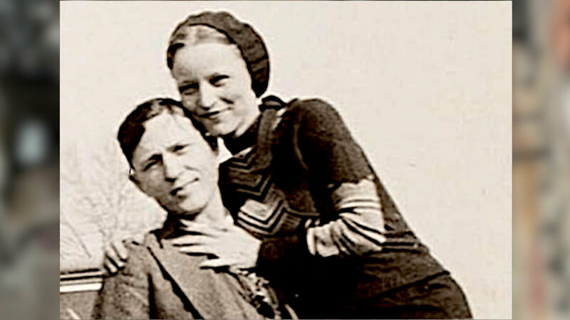 A sepia-toned photograph of the famous outlaws Bonnie and Clyde. The image shows Bonnie Parker leaning over Clyde Barrow, who is seated. Both faces are blurred out. Bonnie is wearing a dark dress with a light-colored scarf around her neck and a beret on her head. Clyde is dressed in a suit with a tie. The background appears to be outdoors, possibly near a car, as part of the vehicle can be seen on the left side of the image. This image captures two notorious figures from American history during the Great Depression era, known for their criminal activities and dramatic end.