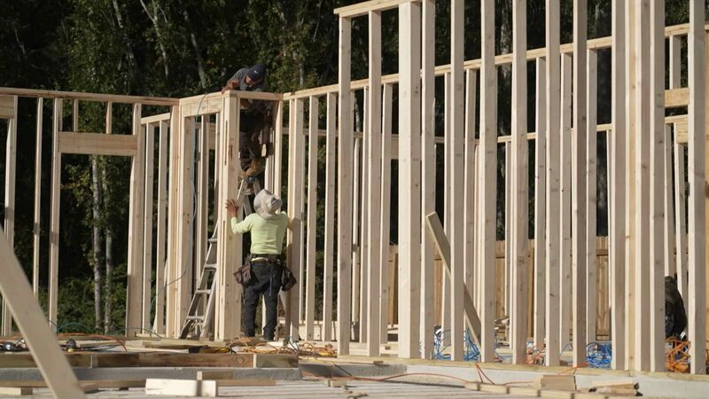 Two construction workers are building a wooden frame structure. One worker is standing on a ladder while the other is on the ground, both working on erecting vertical wooden beams. The background shows trees and construction materials scattered around the site.