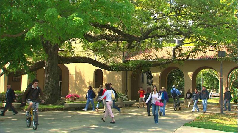 A group of students walk and ride bicycles on a sunny day at Louisiana State University. The scene shows a large tree with sprawling branches providing shade over a pathway in front of an academic building with arched doorways. The students are casually dressed, some carrying backpacks, indicating they are likely heading to or from classes. The image captures the vibrant campus life and the picturesque environment of the university.