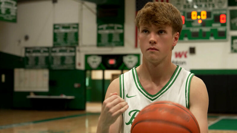 2025 LPB Louisiana Young Hero Mateo Guerrero stands in a gymnasium. He is wearing a white and green basketball jersey with the number partially visible. Guerrero is holding a basketball in the right hand and appears to be preparing to shoot or pass. The background shows a scoreboard displaying '0:00' and various banners on the walls of the gymnasium.