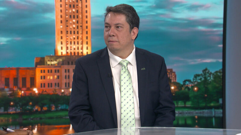Steven Procopio, Policy Director from the Public Affairs Research Council, wearing a dark suit jacket and a light green patterned tie, sits in a studio setting. He has a serious expression and is looking slightly to his left. The background is a digital image of the Louisiana State Capitol building at dusk.