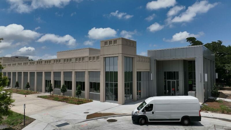 An elevated, wide shot of a modern, two-story building under a blue sky with puffy white clouds. The building has a long, tan concrete section with many windows and a newer section with gray metal siding and a large glass entrance. A white van is parked in the paved lot in the foreground, and small, newly planted trees line the front of the building.