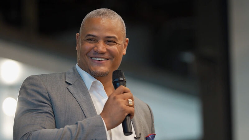 A medium shot of a smiling man with short, graying hair and a goatee, holding a microphone. He is wearing a light gray suit jacket over a white collared shirt and has a red and blue patterned pocket square. The background is softly out of focus.