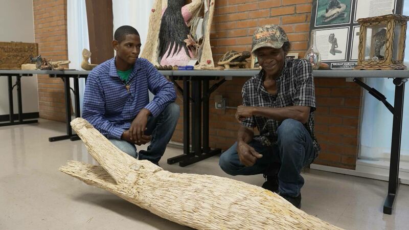 Two men crouch beside a large wooden or woven sculpture on the floor, possibly made of toothpicks or natural fibers. They are in a gallery or exhibit space featuring handcrafted artwork displayed on folding tables against a brick wall. One man wears a plaid shirt and camo hat, the other a blue checkered shirt with a necklace. Visible behind them are sculptures, framed art, and informational signage, highlighting cultural or folk art craftsmanship.